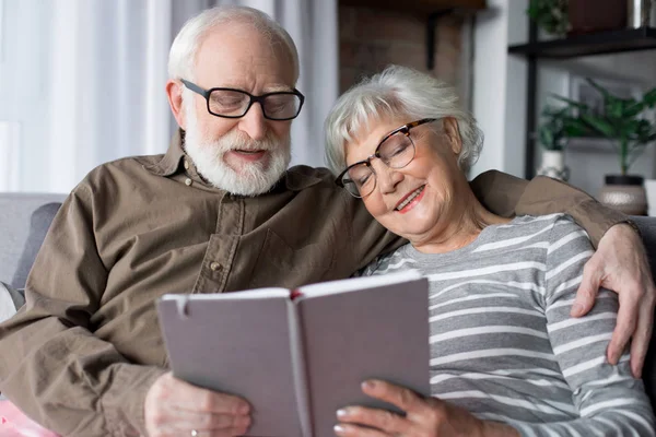 Encantadora pareja de ancianos relajarse en el sofá con libro —  Fotos de Stock