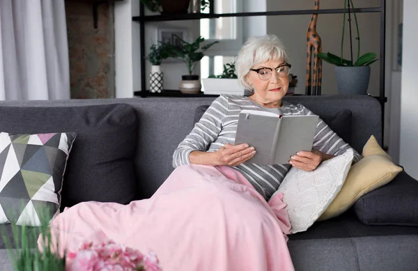 Літня пенсіонерка розслабляється на дивані з книгою — стокове фото