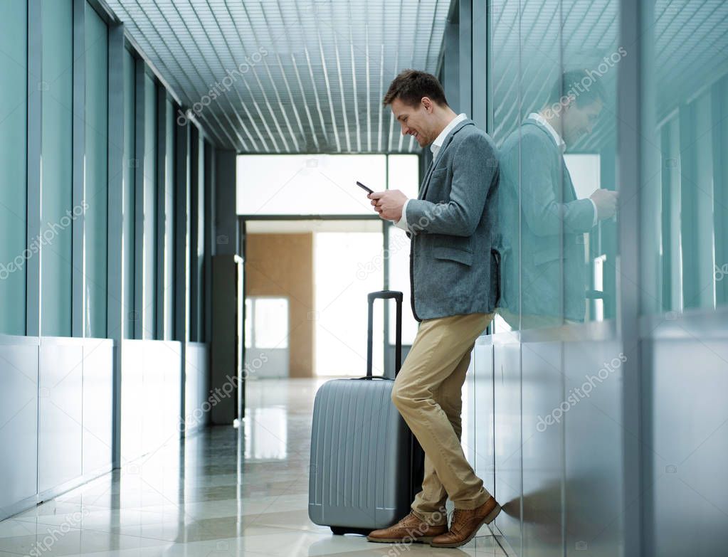 Cheerful guy is standing with suitcase and using modern smartphone