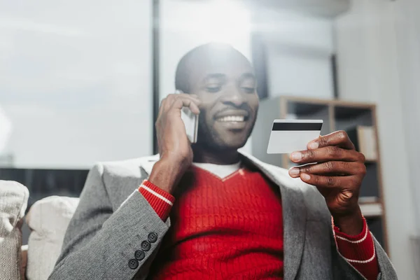 Homem encantado conversando por celular e examinando o cartão bancário — Fotografia de Stock
