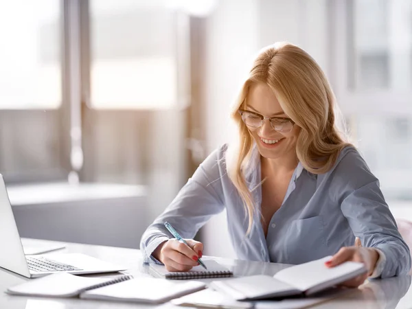 Joyeuse femme blonde écrivant dans un cahier sur le lieu de travail — Photo