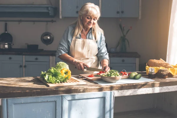 Fröhliche Seniorin kocht Salat mit Musik — Stockfoto