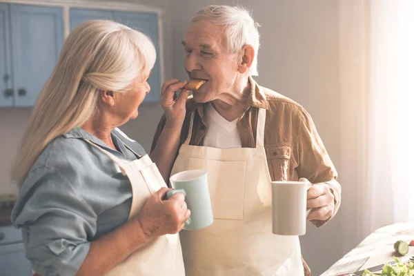 Happy senior couple drinking hot beverage with sweets at home — Stock Photo, Image