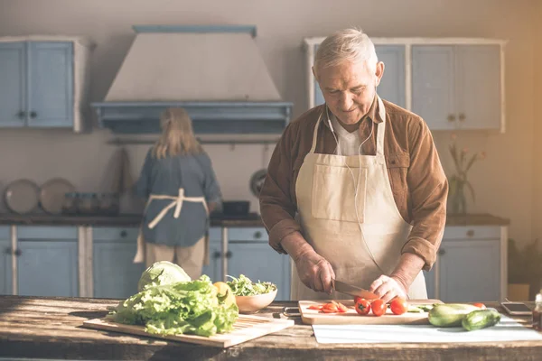 Glücklicher Senior schneidet Gemüse neben Frau in Küche — Stockfoto