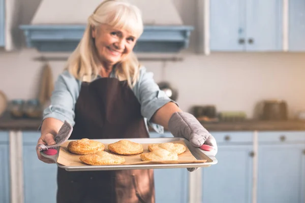 Frohe Seniorin bietet selbstgebackene Brötchen auf Blech an — Stockfoto