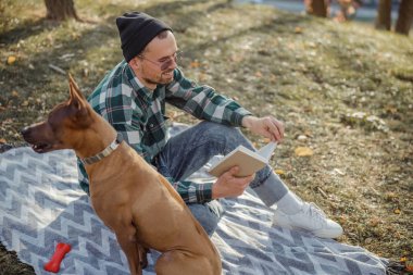 Evcil hayvan sahibi köpekle ormanda kitap okuyor.