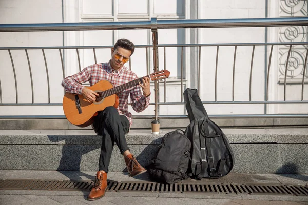 Sorrindo jovem cara está sentado na rua com guitarra — Fotografia de Stock