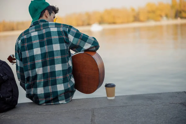 Jovem está sentado perto do rio com guitarra — Fotografia de Stock