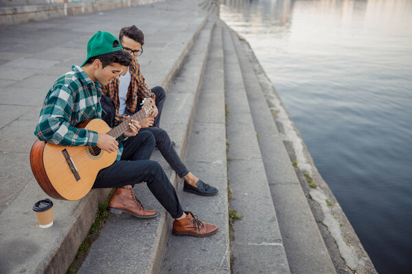 Two young guys sitting near river with guitar