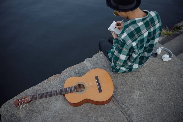 Il chitarrista sta scrivendo sul taccuino vicino al fiume — Foto Stock
