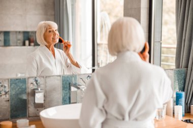 Neşeli yaşlı kadın aynaya bakıyor ve saçlarını tarıyor.