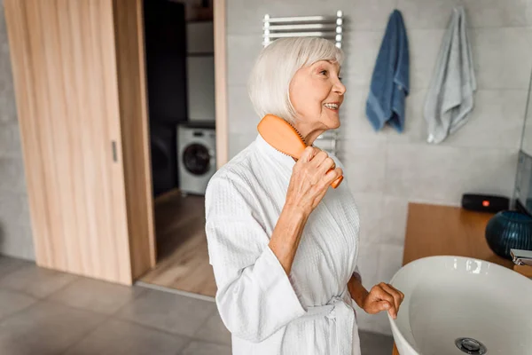 Vrolijke oude vrouw borstelen haar en glimlachen — Stockfoto