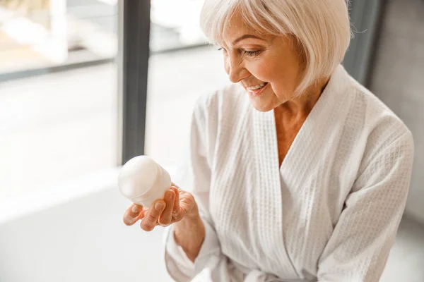 Preciosa anciana mirando crema cosmética y sonriendo — Foto de Stock