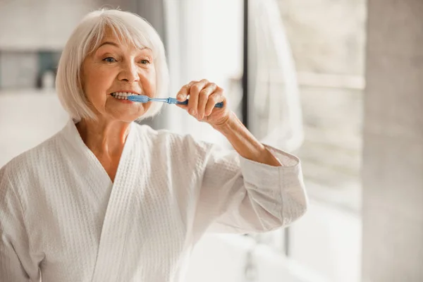 Mujer mayor en albornoz cepillándose los dientes por la mañana — Foto de Stock