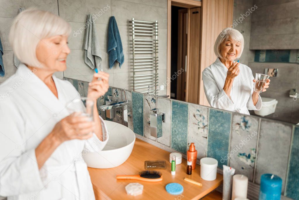 Cheerful old woman standing by the mirror and taking pills