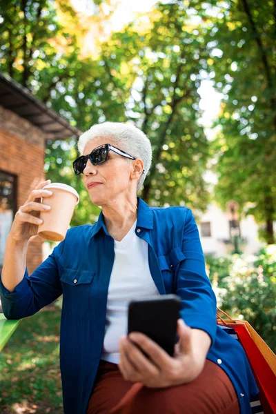 Старшая женщина пьет кофе и использует мобильный телефон на открытом воздухе — стоковое фото