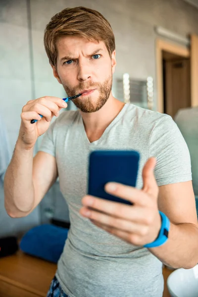 Skäggig ung man borsta tänderna och hålla mobiltelefon — Stockfoto