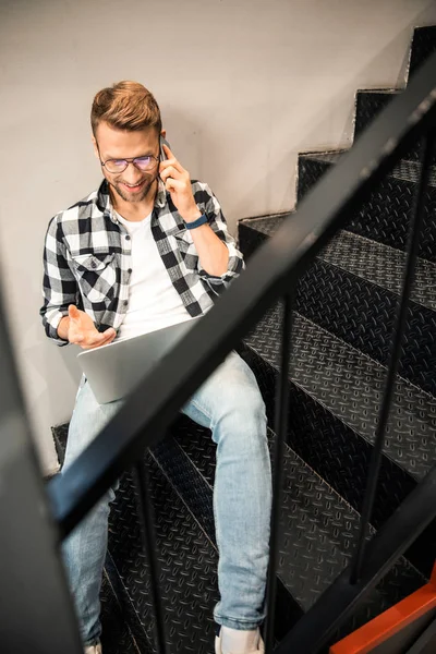 Gülümseyen genç adam merdivenlerde oturuyor ve akıllı telefonda konuşuyor. — Stok fotoğraf