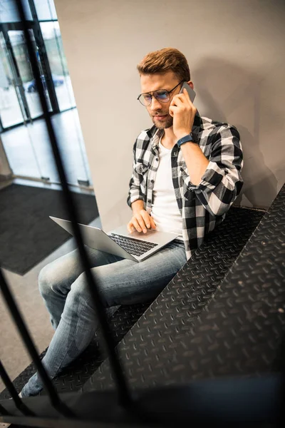 Genç adam dizüstü bilgisayara bakıyor ve akıllı telefondan konuşuyor. — Stok fotoğraf
