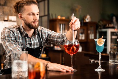 Sakallı barmen tezgahta içki hazırlıyor ve bakıyor.