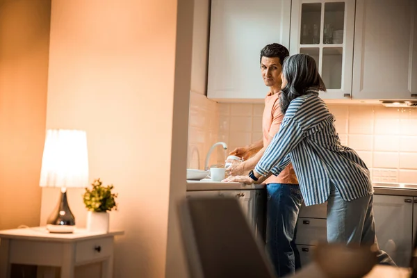 Χαμογελώντας άντρας κοιτάζοντας γυναίκα ενώ πλένει τα πιάτα — Φωτογραφία Αρχείου