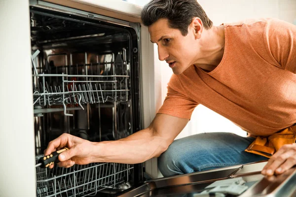 El hombre está utilizando destornillador para reparar el lavavajillas en la cocina —  Fotos de Stock