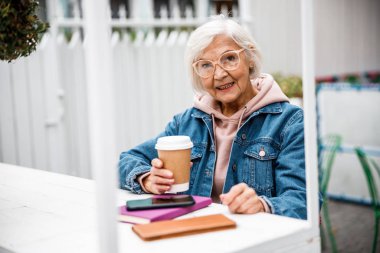 Elinde telefon ve kahve dükkanı fotoğrafı olan gülümseyen yaşlı bayan.