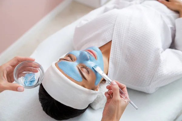 Kosmetikerin trägt blaue Maske auf Gesicht der Dame auf — Stockfoto