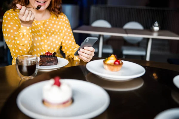 Señora sonriente comiendo pastel y usando teléfono inteligente —  Fotos de Stock