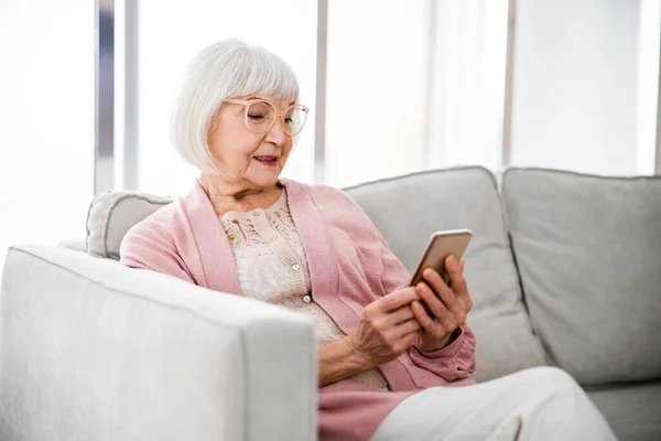 Усміхнена стара жінка використовує смартфон вдома — стокове фото
