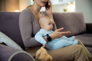 Gülümseyen genç anne bebekle oturuyor ve akıllı telefon kullanıyor.