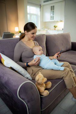 Mutlu genç anne çocukla oturuyor ve cep telefonu kullanıyor.