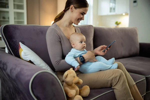Usmívající se mladá máma sedí s dítětem a drží smartphone — Stock fotografie