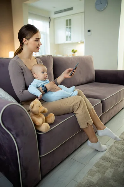 Sorrindo jovem mãe sentada com o bebê e usando smartphone — Fotografia de Stock