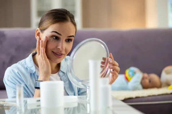 Mãe bonita feliz usando creme facial no quarto — Fotografia de Stock