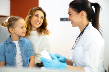 Küçük çocuk annesinin dizlerinin üstüne çöküp doktoru dinliyor.