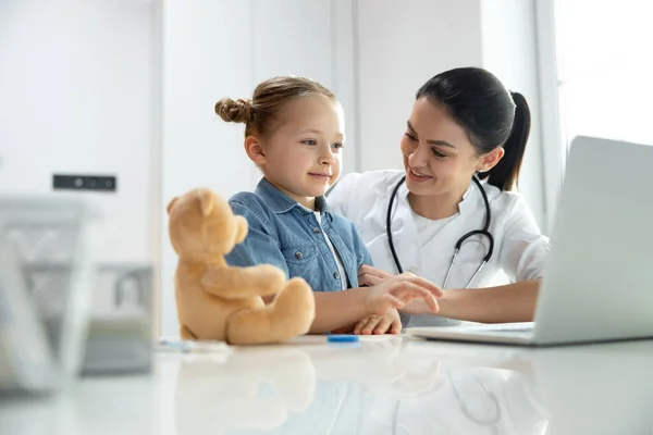 Amable pediatra sonriendo a su pequeña y linda paciente en el interior —  Fotos de Stock