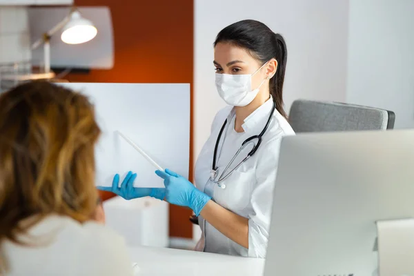 Серйозна жінка-лікар в медичній масці, що показує щось на аркуші паперу в лікарні — стокове фото