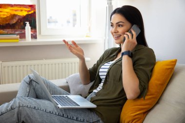 Güzel bayan dizüstü bilgisayar kullanıyor ve evde telefon görüşmesi yapıyor.