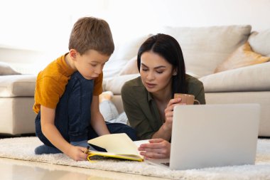 Anne ve oğlu dizüstü bilgisayarı ve not defteriyle halı fotoğrafında.