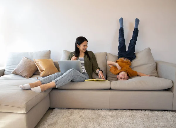 Laughing woman with laptop watching her son lying upside down — Stock Photo, Image