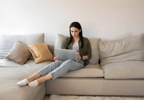 Mujer joven seria sentada en el sofá con el dispositivo moderno foto de stock — Foto de Stock