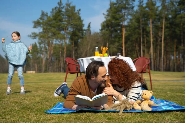 Liefdevol getrouwd stel liggend op deken op groen gras en leesboek — Stockfoto