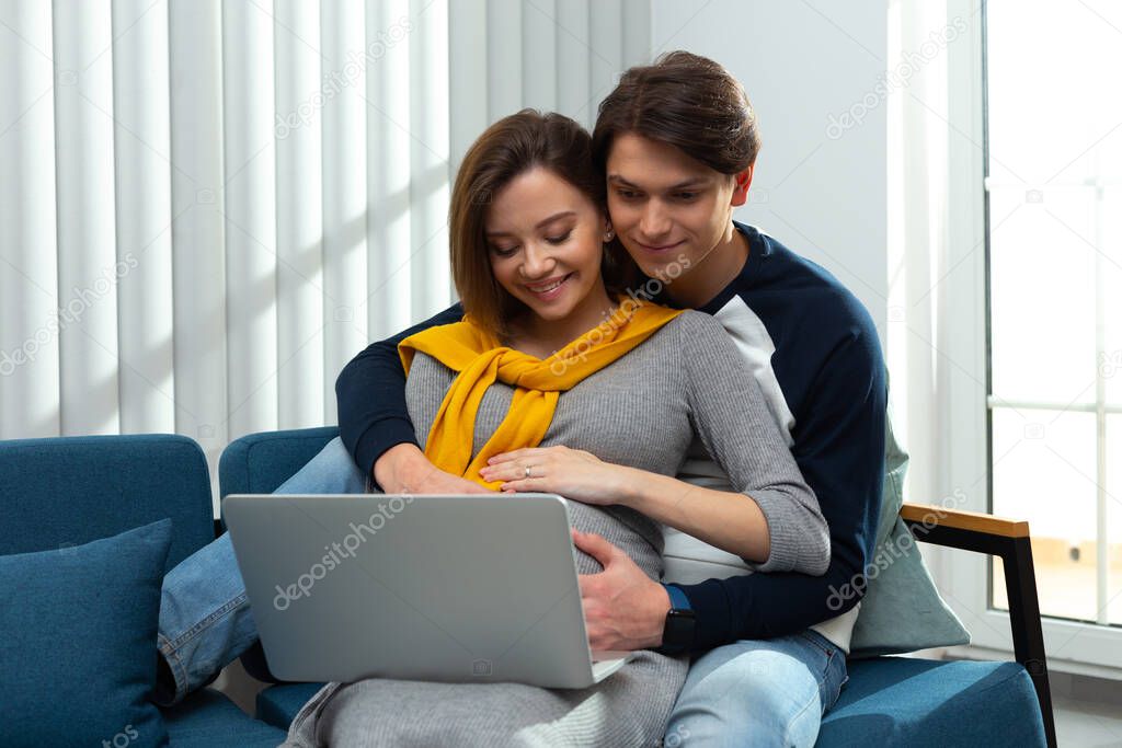 Mirthful pregnant couple at home with modern gadget