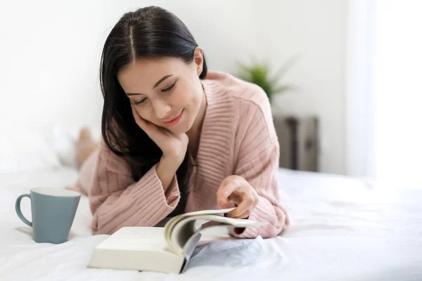 Νεαρή γυναίκα χαλαρωτικό και διαβάζοντας το βιβλίο στο κρεβάτι στο σπίτι — Φωτογραφία Αρχείου