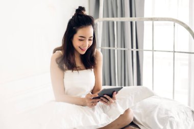 Mutlu Asyalı kadın yatakta tablet bilgisayar kullanarak rahatlıyor.