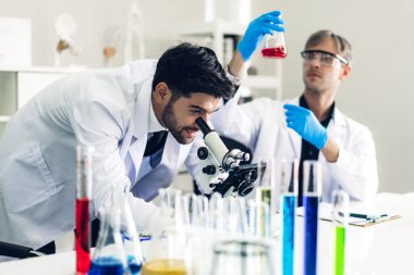 Profesyonel iki bilim adamı araştırma yaparken kimyasal bir deney yapıyor ve mikroskobu kullanarak modern laboratuvar deney tüpünde kimyasalları karıştırıyor.