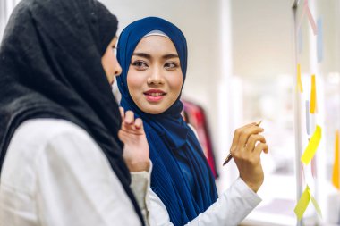 Yaratıcı, mutlu, mutlu, gülümseyen iki kolej. İki Müslüman iş kadını strateji analizi ve beyin fırtınası yapıyor. Ofisteki cam pencere projesine başlama projesini paylaşmak için çıkartma yapıştırıyor.