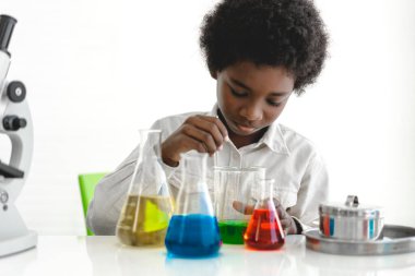 Afrika kökenli, sevimli, küçük bir çocuk araştırma öğreniyor ve kimyasal bir deney yaparken fen bilimleri sınıfında sıvıyı analiz edip karıştırıyor. Eğitim ve bilim konsepti.