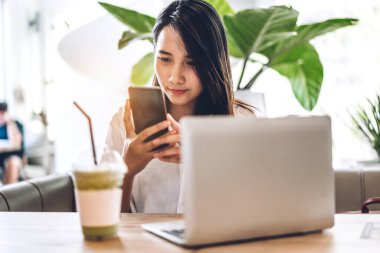 Gülümseyen mutlu Asyalı bir kadının portresi akıllı telefon teknolojisi kullanarak rahatlıyor ve sandalyede otururken çalışıyor. Kafede ve restoranda dizüstü bilgisayar kullanan genç hippi kız serbest çalışıyor.